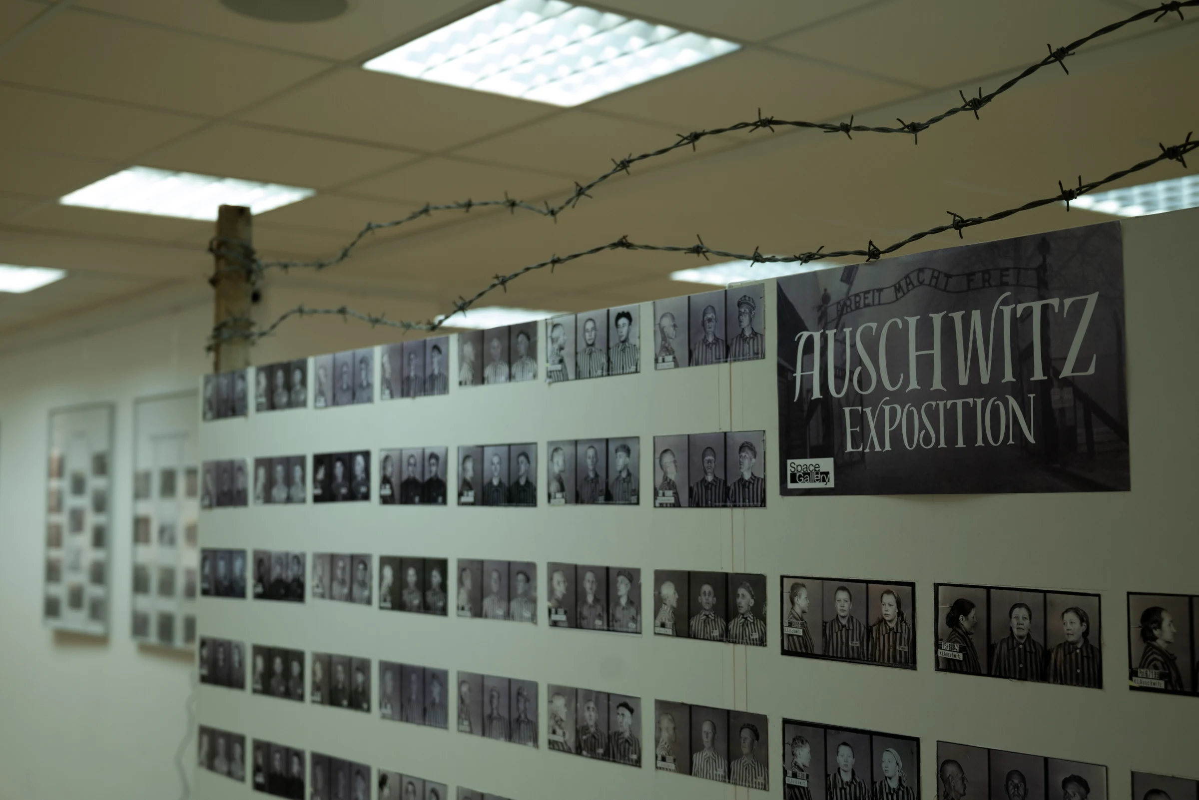 Výstavní stěna s portréty vězňů, ostnatým drátem a názvem Auschwitz Exposition